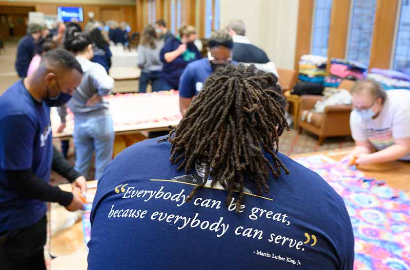 A group of people in a room work at tables. A Black man with dreads is in focus in the foreground with his back facing the camera. His shirt says 'Everybody can be great, because everybody can serve.'