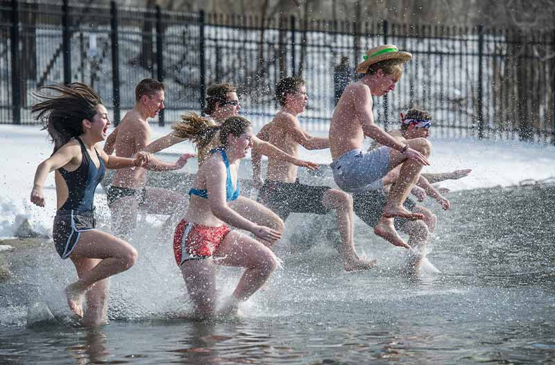 A group of seven people wearing bathingsuits running into a lake. The grass behind them is covered in snow.