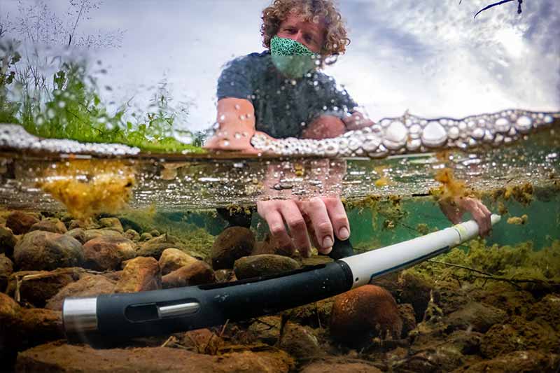 A man wearing a mask sticks an instrament under shallow water.