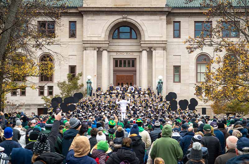 The Notre Dame Marching Band performs outside on the steps of Bond Hall in front of a group of fans.