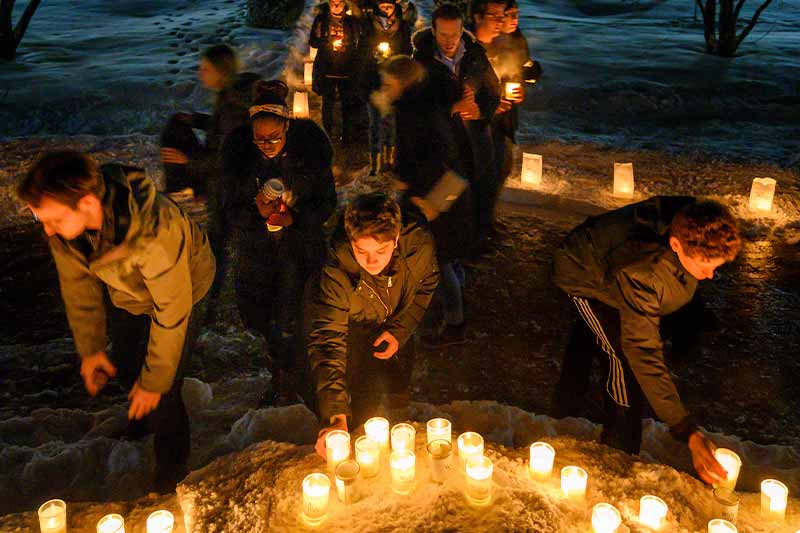 At night people wearing coats place lit candles on the ground during a prayer service.