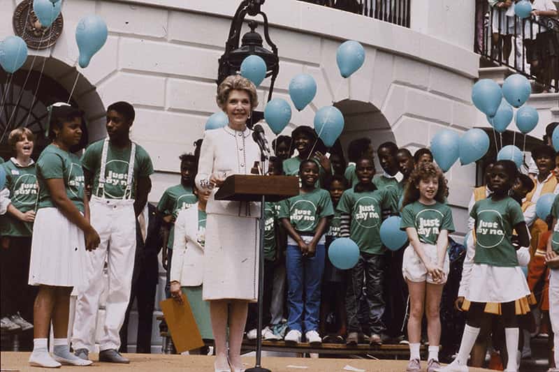 Nancy Reagan stands in front of a podiam as young children stand behind her in green t-shirts that say “Just Say No”.