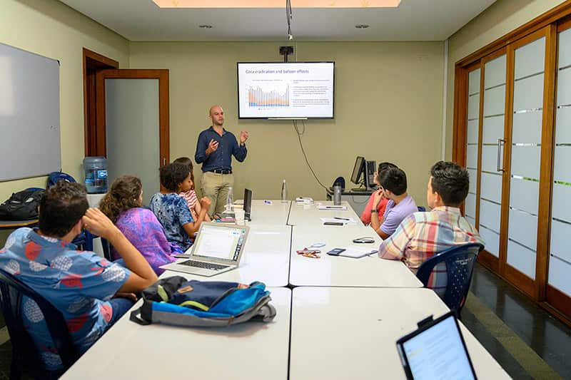A man stands next to a large monitor and presents to a group of people sitting down at a table.