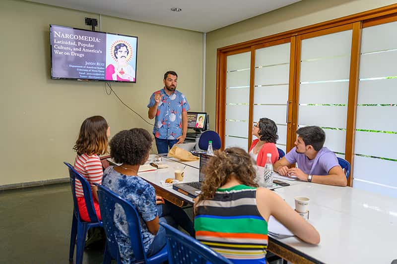 A man stands next to a large monitor and presents to a group of people sitting down at a table.