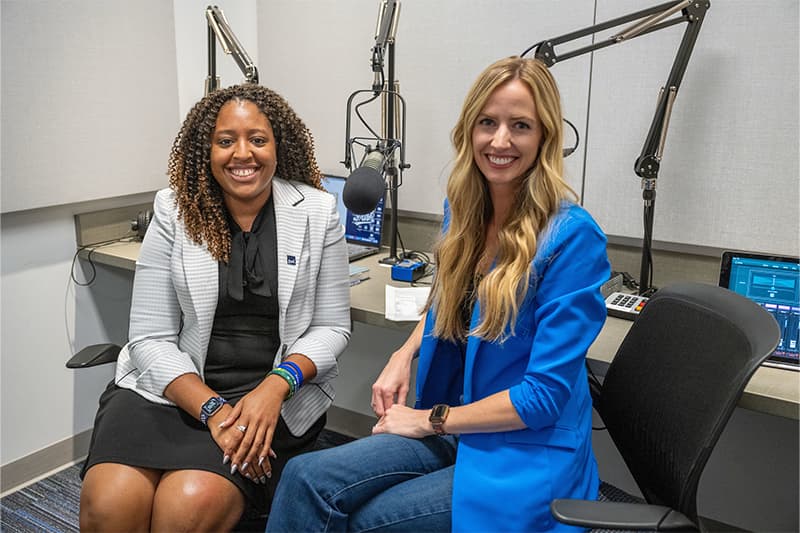 Internal Communications Director Jenna Liberto sits to the right of JP Abercrumbie, executive associate athletics director for culture and engagement. Both women pose smiling with recording mics in the background