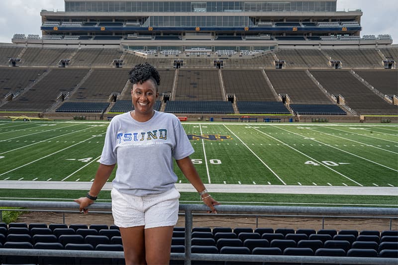 South Bend native Jennifer V. Liddell stands smiling in front of Notre Dame Stadium's 50-yard line wearing a TSU-UND game t-shirt.