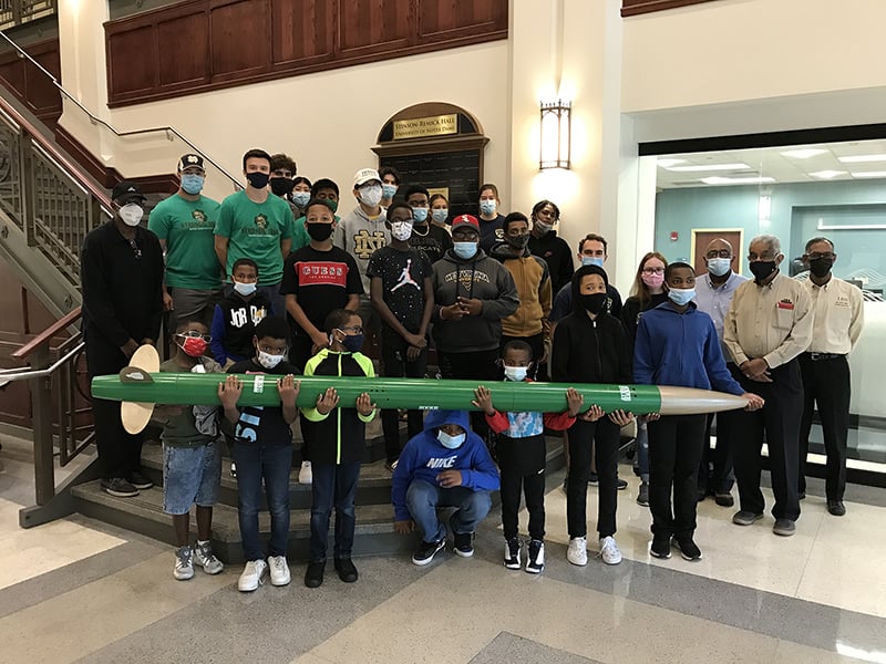 Mentors and mentees from 100 Black Men stand at the base of a staircase holding a rocket.