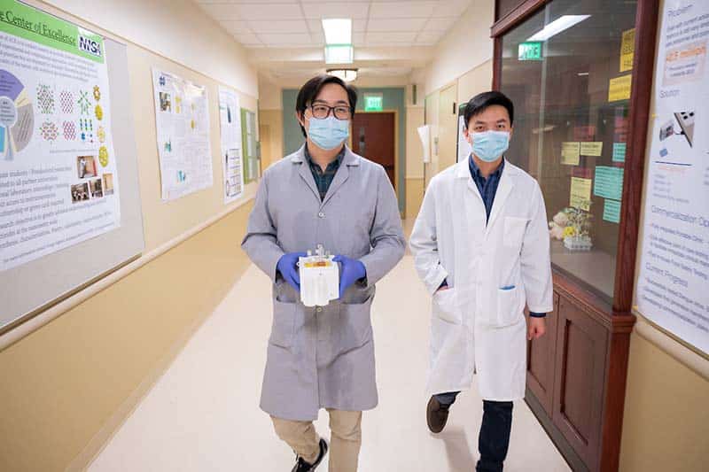 Two men in lab coats and face masks walk down a hallway carrying a white box, the testing device.