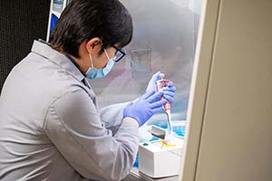 A scientist with mask and gloves and lab coat conducts testing.