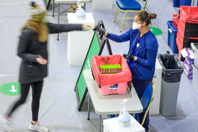 A blurred student hands a saliva sample to a nurse wearing scrubs and mask at collection station.