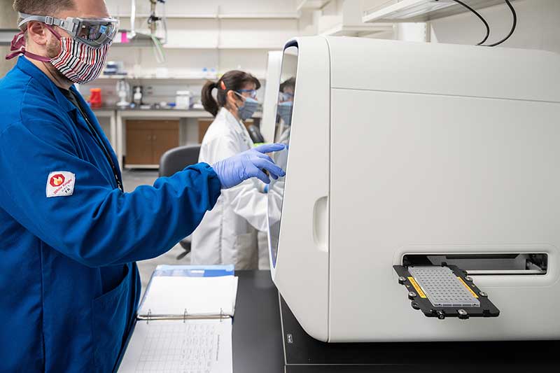 A man in a lab wearing a mask, safety glasses, lab coat, and latex gloves works with a PCR machine.
