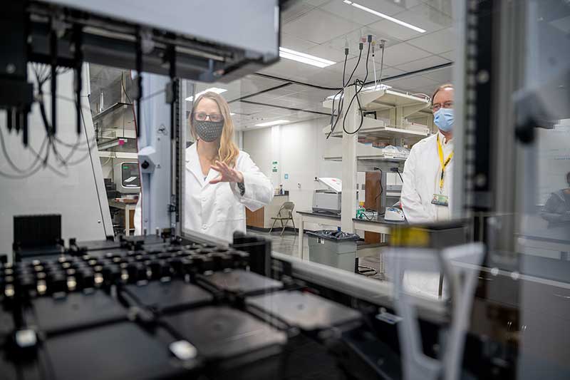 A woman wearing a mask stands behind a piece of lab equipment.