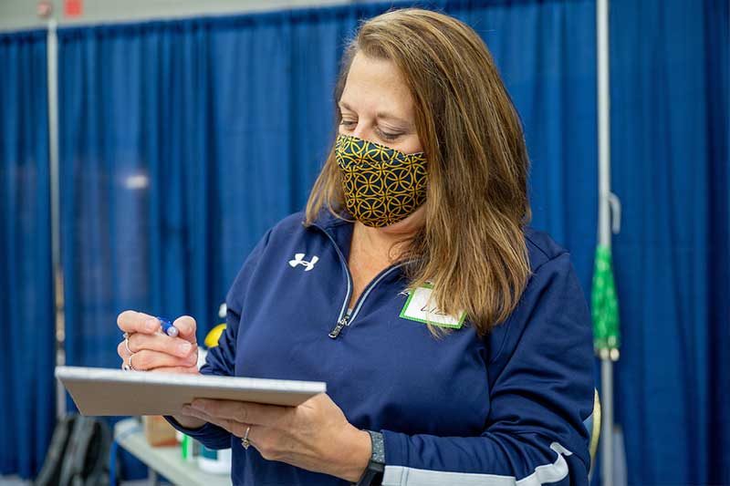 A masked employee of the University Testing Center jots down notes on a notepad.