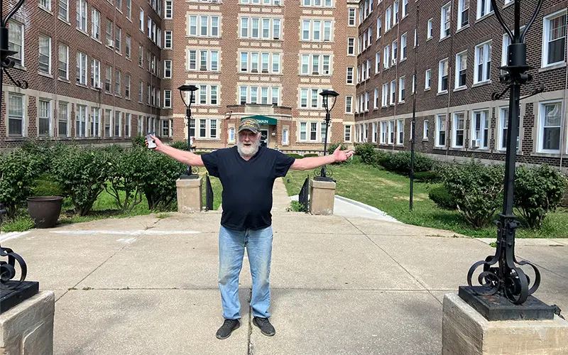 Jack West, a white man, with a white beard stands in front of a brown-bricked apartment building with his arms wide open.