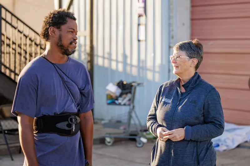 Margie Pfeil stands with a black man in front of an industrial looking building. There's a full shopping cart in the background.