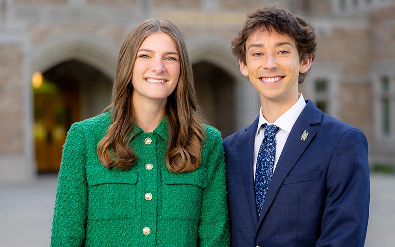 Two smiling students stand in front of an arched entryway to a Notre Dame building. The woman on the left wears a green jacket, and the man on the right wears a navy suit with a floral tie.