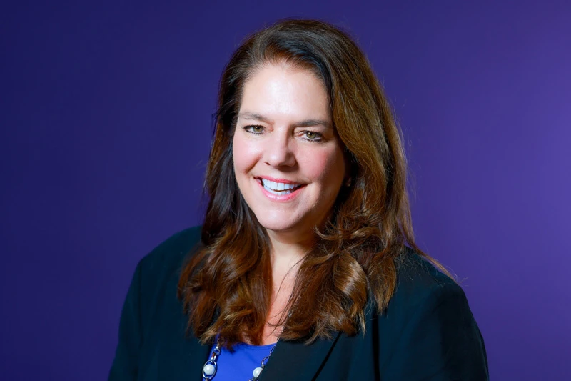A woman with shoulder-length brown hair smiles against a purple backdrop. She wears a black blazer over a royal blue top and a silver necklace.