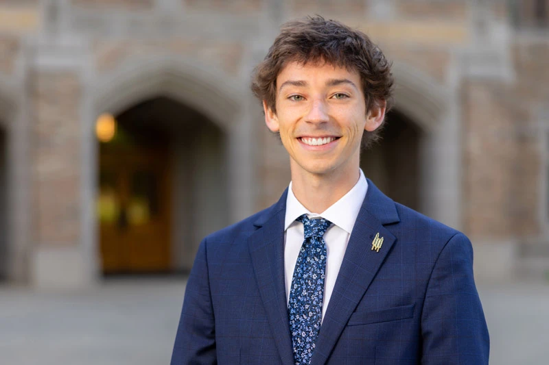 A young white man wearing a navy blue suit, floral tie, and a gold Notre Dame lapel pin smiles in front of an arched entryway of a stone building on campus.