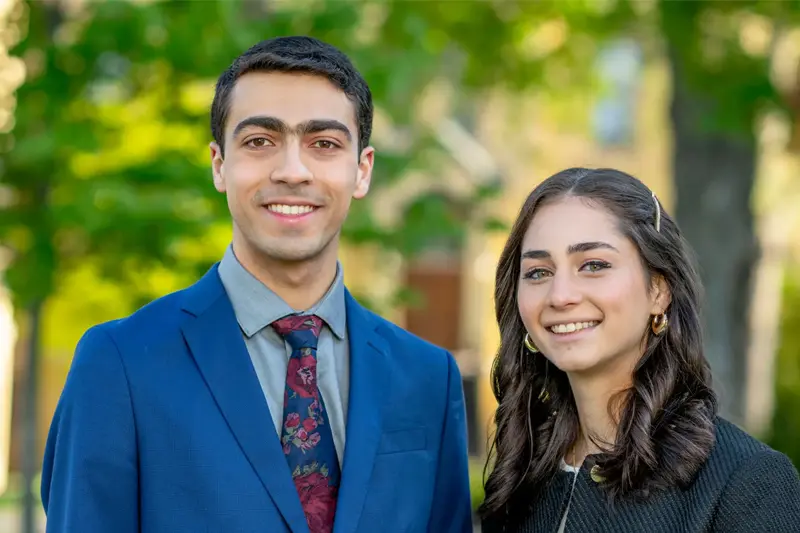 Two students, a man in a blue suit and floral tie and a woman in a black jacket, smile at the camera outdoors on a sunny day.