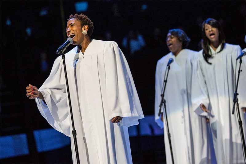 Three women wearing white choir robes sing on stage. Two of the singers are in the background.