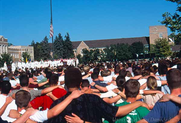 An archive photo of Mass on the South Quad after the attacks. Congregants have their arms around each others shoulders and look towards the altar and the US flag at half-mast.