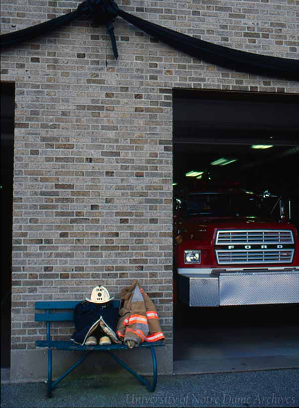 A black bunting hangs above the fire department garage door. A fire truck is inside. Outside on a bench sits a firefighter coat and helmet.