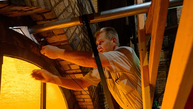 Man feeding wires through a tube protruding through a hole above a stained glass window from the interior of the basilica steeple.