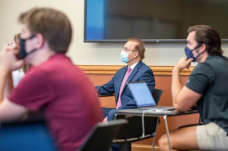 A masked student man and two masked students sit at desks in a classroom.