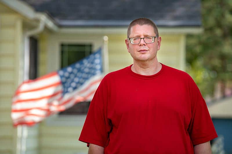 A man wearing a red t-shirt stands in front of a yellow house where an American flag blows in the wind.