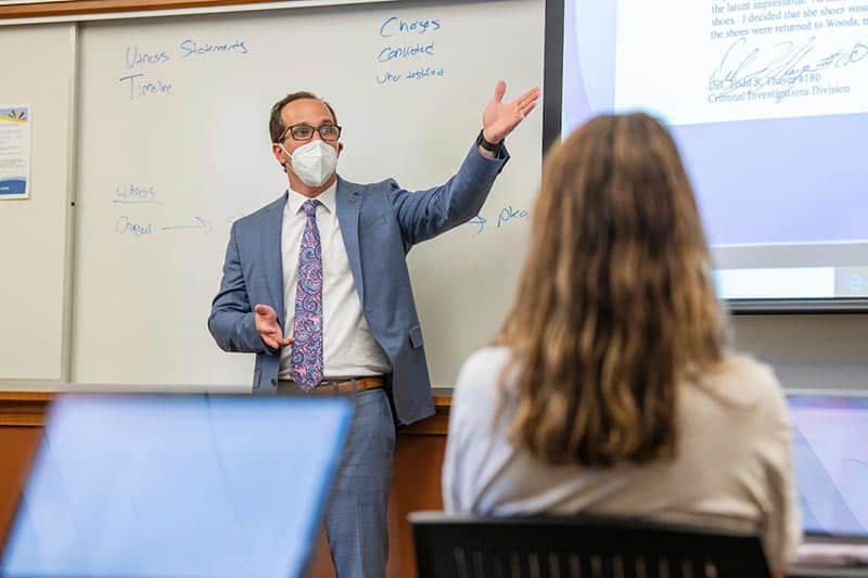 A masked teacher points to a projected screen.