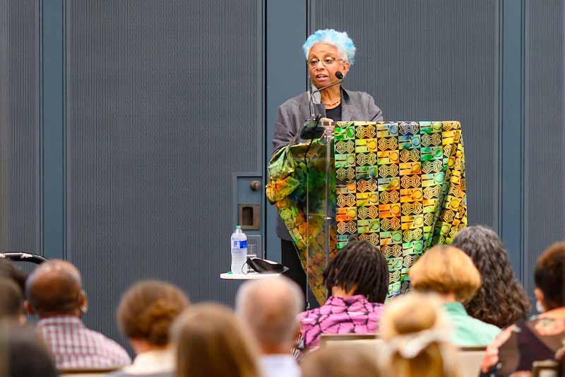 A woman stands at a podium and speaks to a group of people.