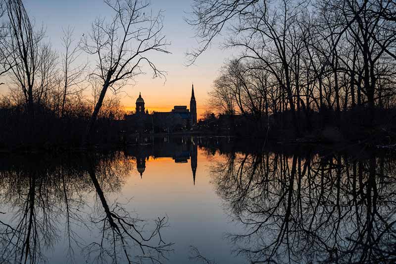 A sunrise over a lake. Notre Dame's main building and Basicila are far in the distance. Bare trees reflect off of the water.