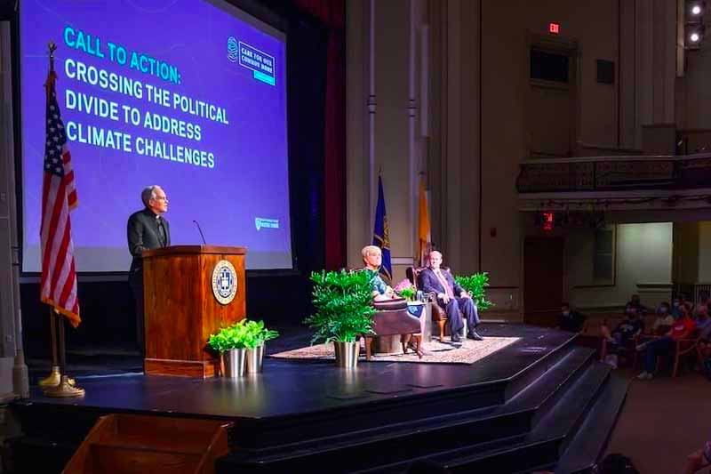 Fr. John stands on stage and at a podium in front of a large screen. Two other individuals sit in chairs.