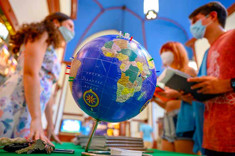 A globe sits on a table, in focus. In the background are three blurred out people talking, each wearing masks.