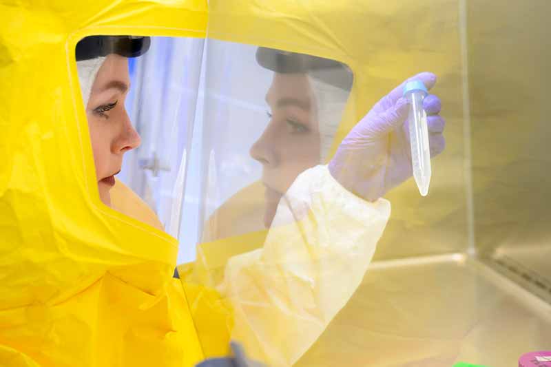 A woman wearing a yellow hazmat suit and gloves holds up a test tube.