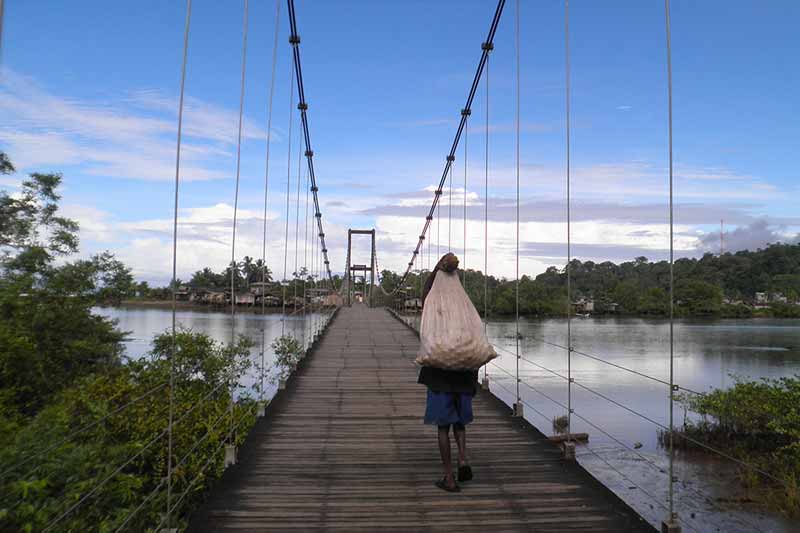 A young child holds a bag over their back and walks on a bridge  over a body of water.