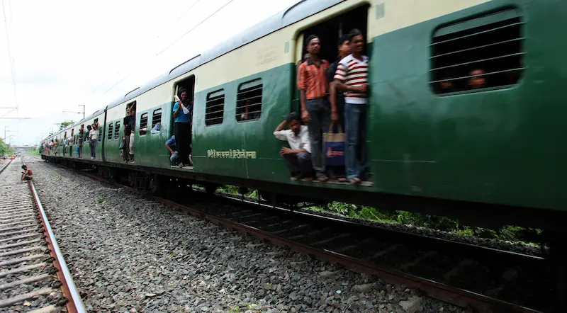 A crowded green and off-white train moves along tracks, with people hanging out of the open doorways and windows.