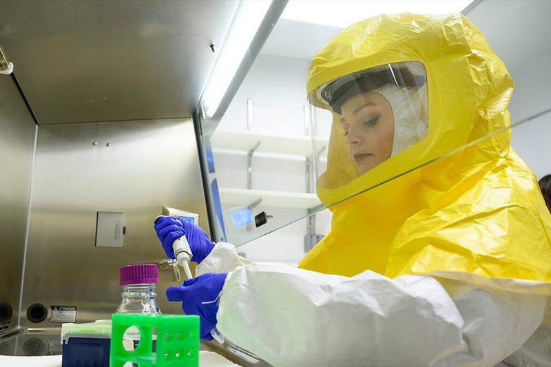 Research staff member withdrawing fluid from vial while dressed in a yellow biosafety suit.