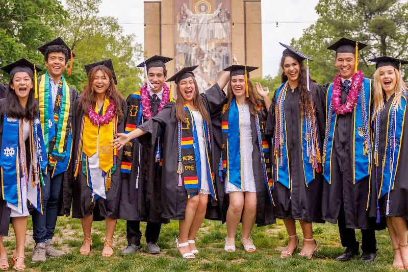 Nine men and women wearing graduation robes and caps smile arm in arm in front of Hesburgh Library at the University of Notre Dame.