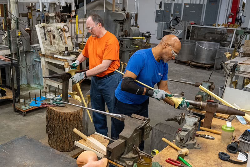 Two men shaping the trumpets by hand with anvils and hammers in a workshop.