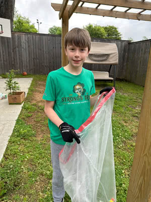 A boy stands in a backyard wearing yard gloves and is holding a trash bag.