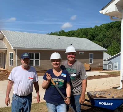 Three people wearing hard hats stand in a yard. One is holding a shovel.