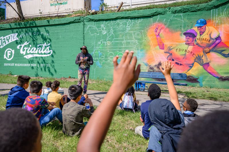 A man standing in front of a brightly painted mural talking to a group of students.