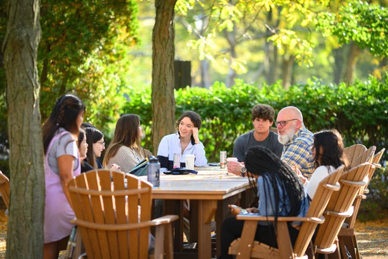 A group of students and a professor sit outside at a wooden table underneath green trees at the LaFortune student center.