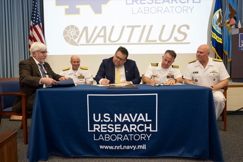 Jeff Rhoads sits at a table with a man and three navy officers while signing a piece of paper.