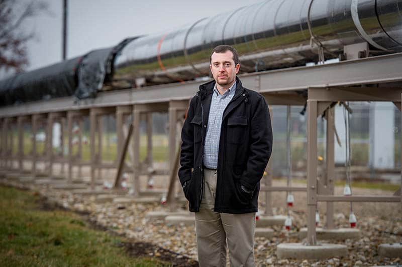 A man stands in front of a long large silver tube, a wind tunnel.