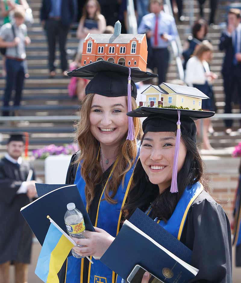 Two graduates smile during commencement holding diplomas and wearing decorated graduation caps. One cap features a model Habitat home and the other Notre Dame's South Dining Hall.