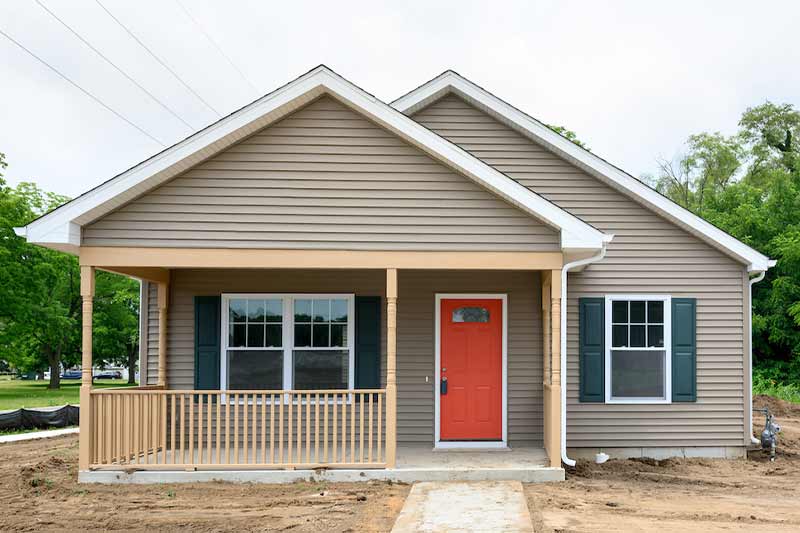 A newly built Craftsman Bungalow home with a covered porch, light brown siding and a bright blood-orange door. The home is surrounded by dirt.