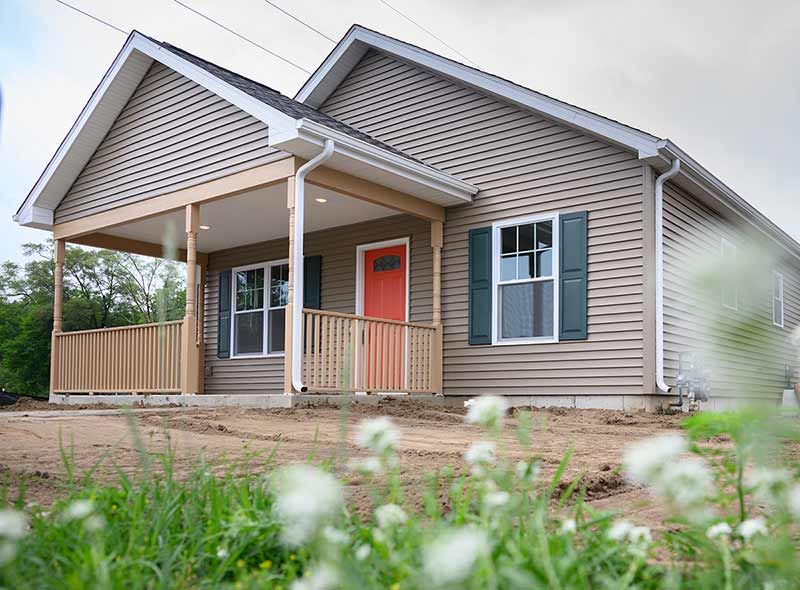 A newly built Craftsman Bungalow home with light brown siding and a bright blood-orange door. The home is surrounded by dirt and in the foreground is blurred out grass and white flowers.