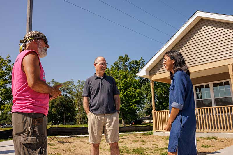 Three individuals talk while standing outside in front of a newly build home. Behind them is a clear blue sky and green trees that line the background.